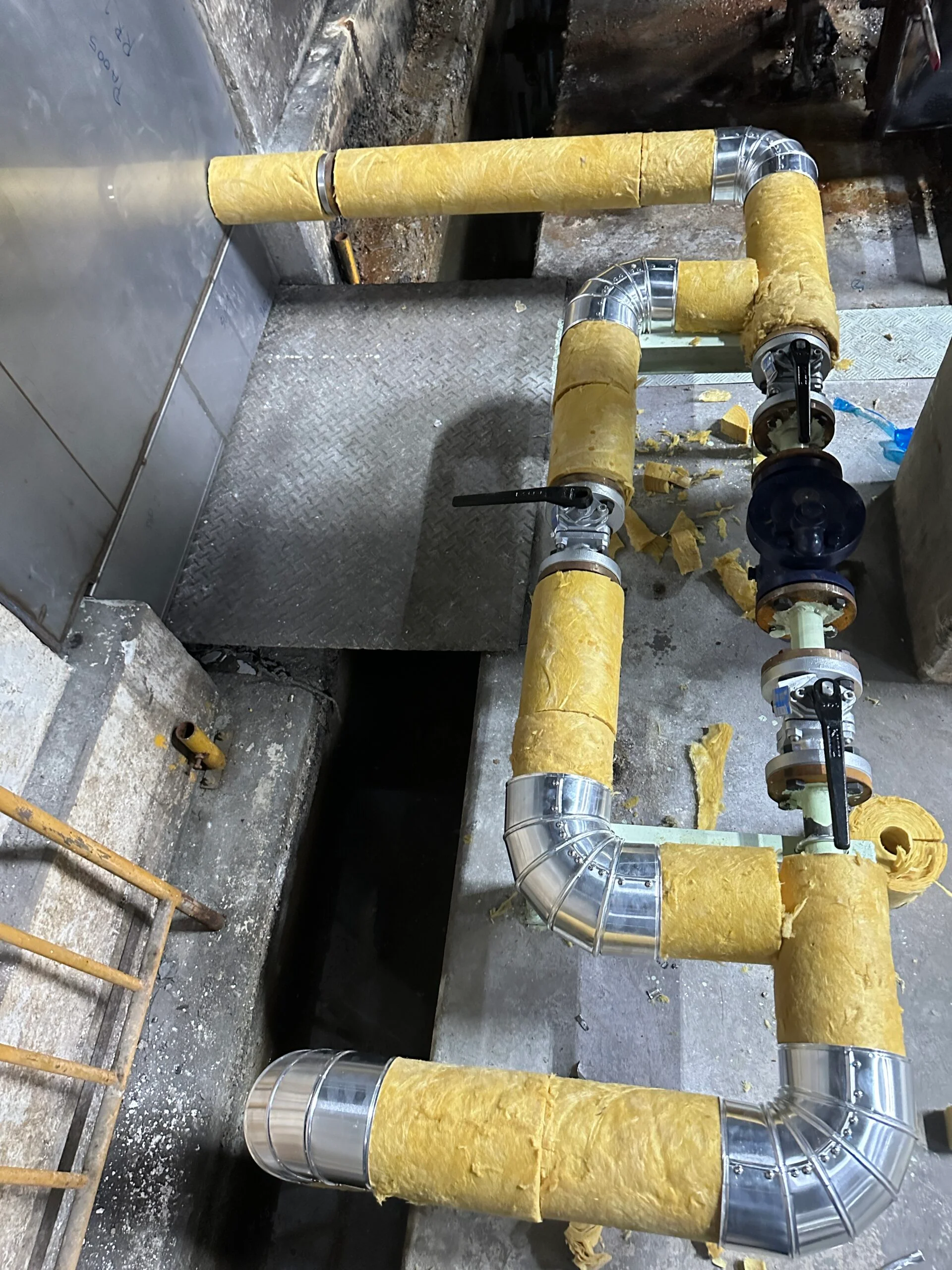 Yellow-insulated industrial pipes with silver elbows and black valves run over a metal floor above open trenches in a work area