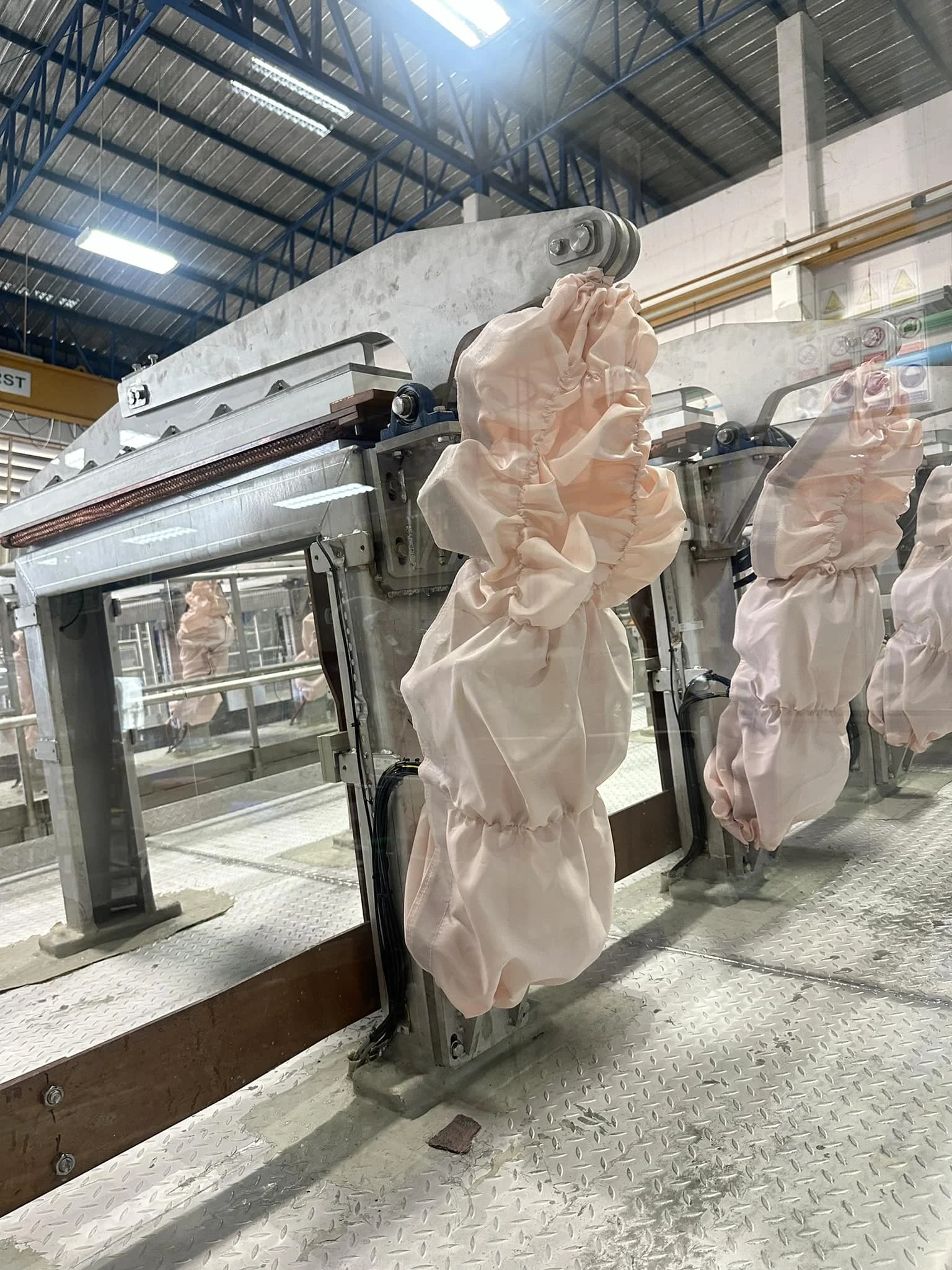 Conveyor system with pink cloth bundles hanging, inside an industrial setting with metal beams and patterned steel flooring. Bright lighting.