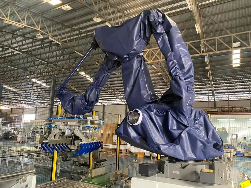 Industrial robot arm covered in blue tarp, operating in a factory with metal roof. The arm has a gripping tool. No text visible.