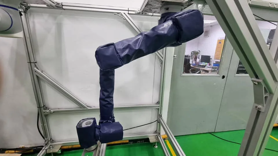 A blue padded robotic arm in a metal frame against a gray industrial background with green flooring and a glass door. Text reads