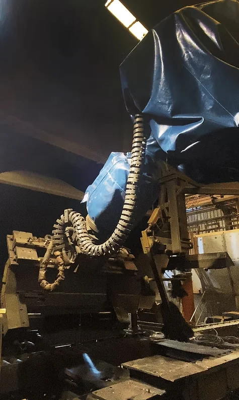 Industrial machine in a dimly lit factory, covered in blue tarp and flexible tubing. Overhead lights cast a moody atmosphere.